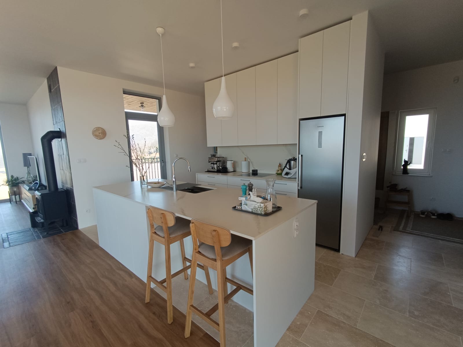 Jasna kuchnia z wyspą, drewnianymi hokerami i białymi szafkami. Widok na zewnątrz przez okno. Nowoczesny design.