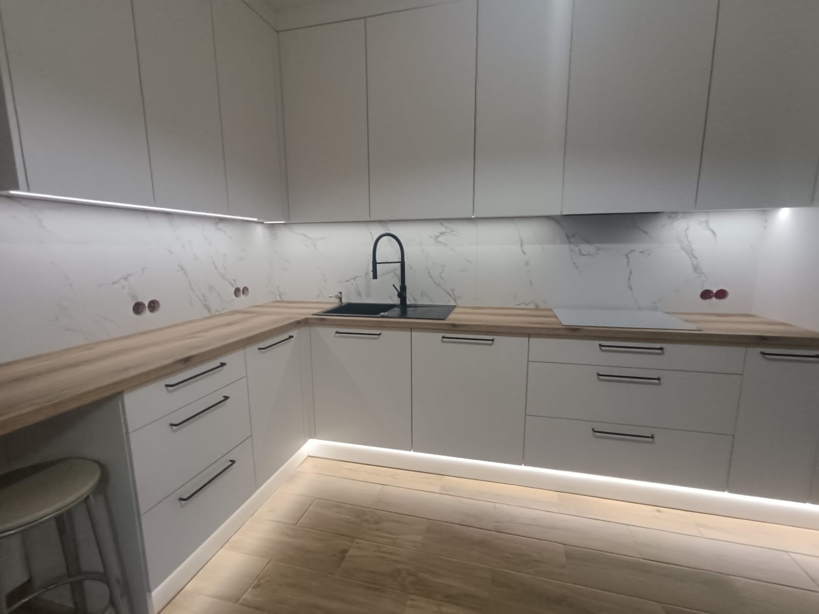 Nowoczesna, jasna kuchnia z drewnianym blatem i marmurowym backsplashiem. Oświetlenie LED pod szafkami dodaje elegancji. Widoczny fragment stołka barowego.