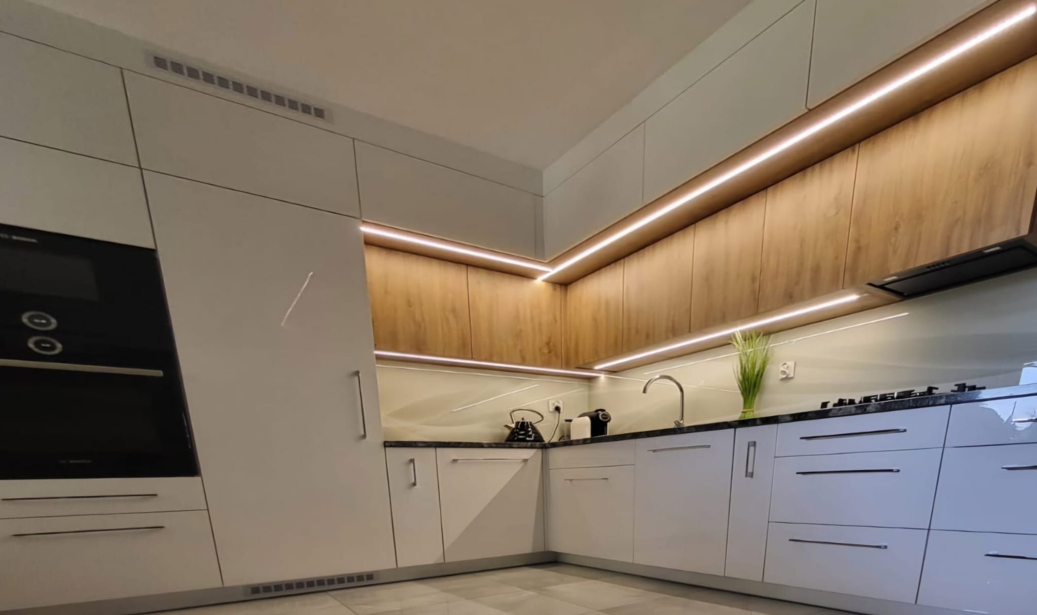 Nowoczesna kuchnia z białymi, lakierowanymi frontami i drewnianymi szafkami górnymi, oświetlona listwami LED. Minimalistyczny design z czarnym blatem i AGD.