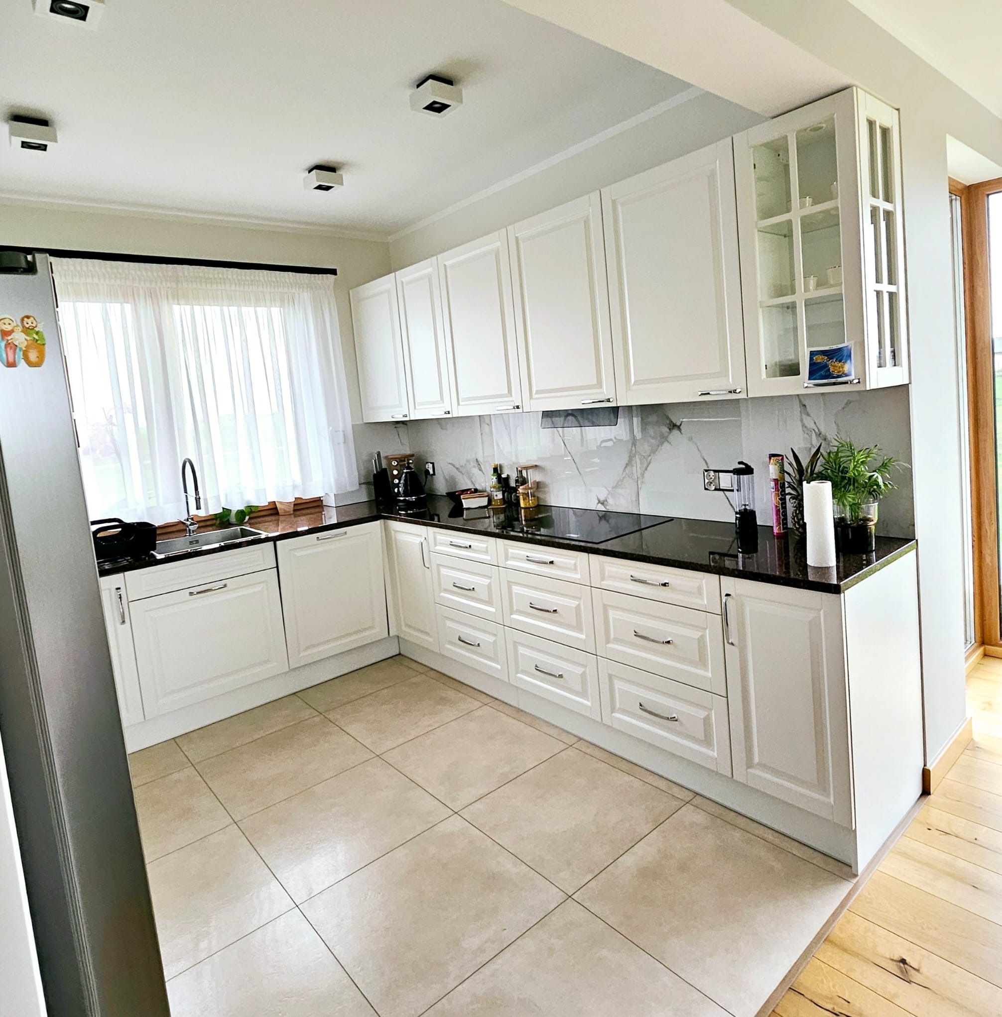 Jasna kuchnia w kształcie litery L z białymi frontami szafek, czarnym blatem i marmurowym backsplashiem. Widoczne okno z firanką i drewniana podłoga w przejściu.