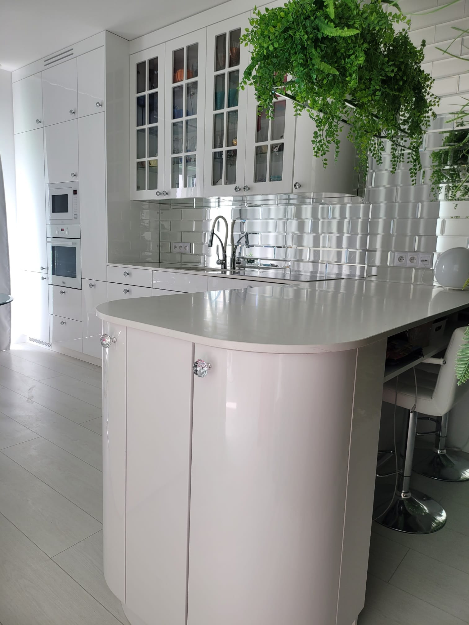 Biała kuchnia na wymiar z wyspą o obłych kształtach i kryształowymi gałkami, wykończona błyszczącymi kaflami i wiszącymi roślinami. Nowoczesny design.