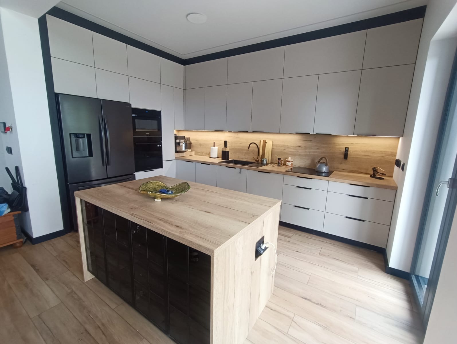 Nowoczesna kuchnia z wyspą: szare szafki z czarnymi uchwytami, drewniany blat, czarny sprzęt AGD i podświetlenie LED. Wyspa z czarnymi frontami.