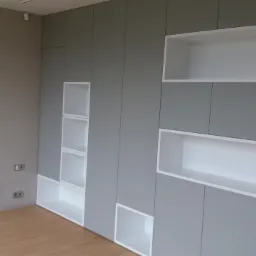 Szara zabudowa ścienna z białymi wnękami i półkami w minimalistycznym wnętrzu z drewnianą podłogą i betonową ścianą.