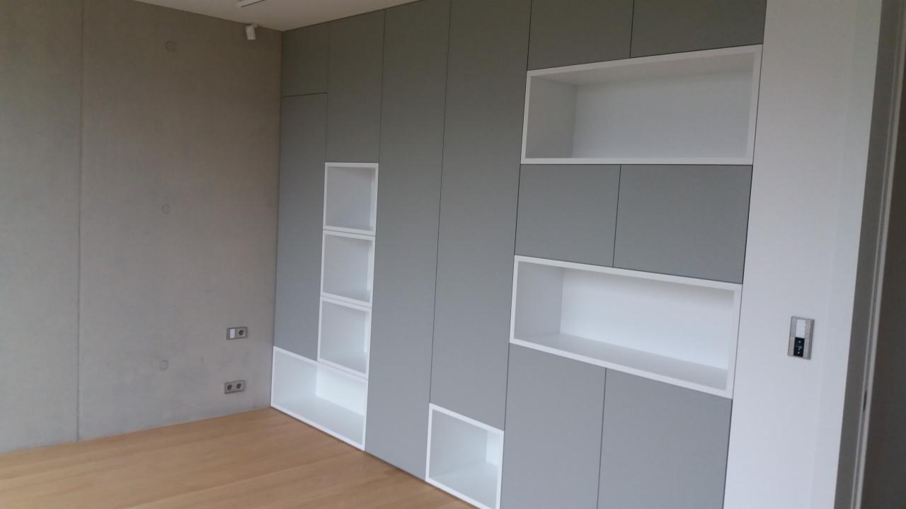 Szara zabudowa ścienna z białymi wnękami i półkami w minimalistycznym wnętrzu z drewnianą podłogą i betonową ścianą.