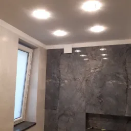 Wykończona łazienka z szarą marmurową okładziną ścian, wnęką z półką, białym oknem z czarnym parapetem i oświetleniem sufitowym punktowym.