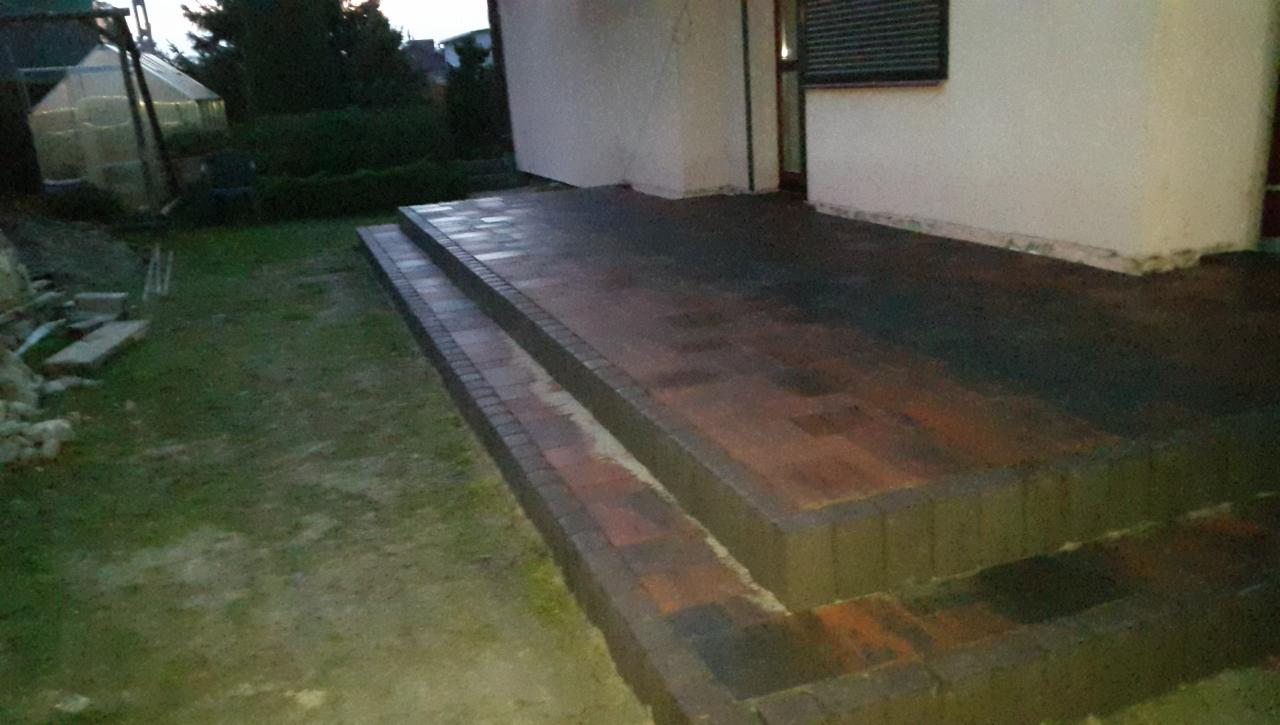 Nowo wyłożony taras z cegły klinkierowej z dwoma stopniami, prowadzący do domu z widoczną szklarnią w tle.