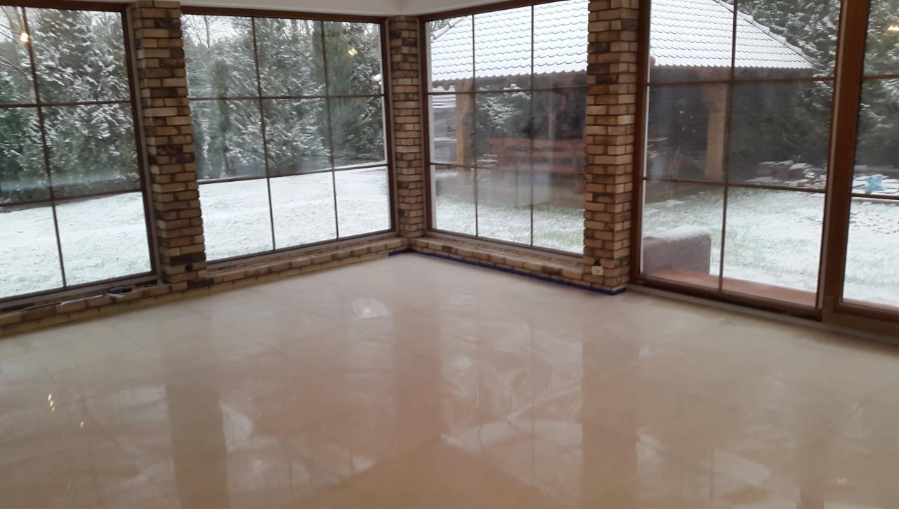 Jasna, błyszcząca podłoga z płytek w oranżerii z ceglanymi filarami i dużymi oknami z widokiem na ośnieżony ogród.