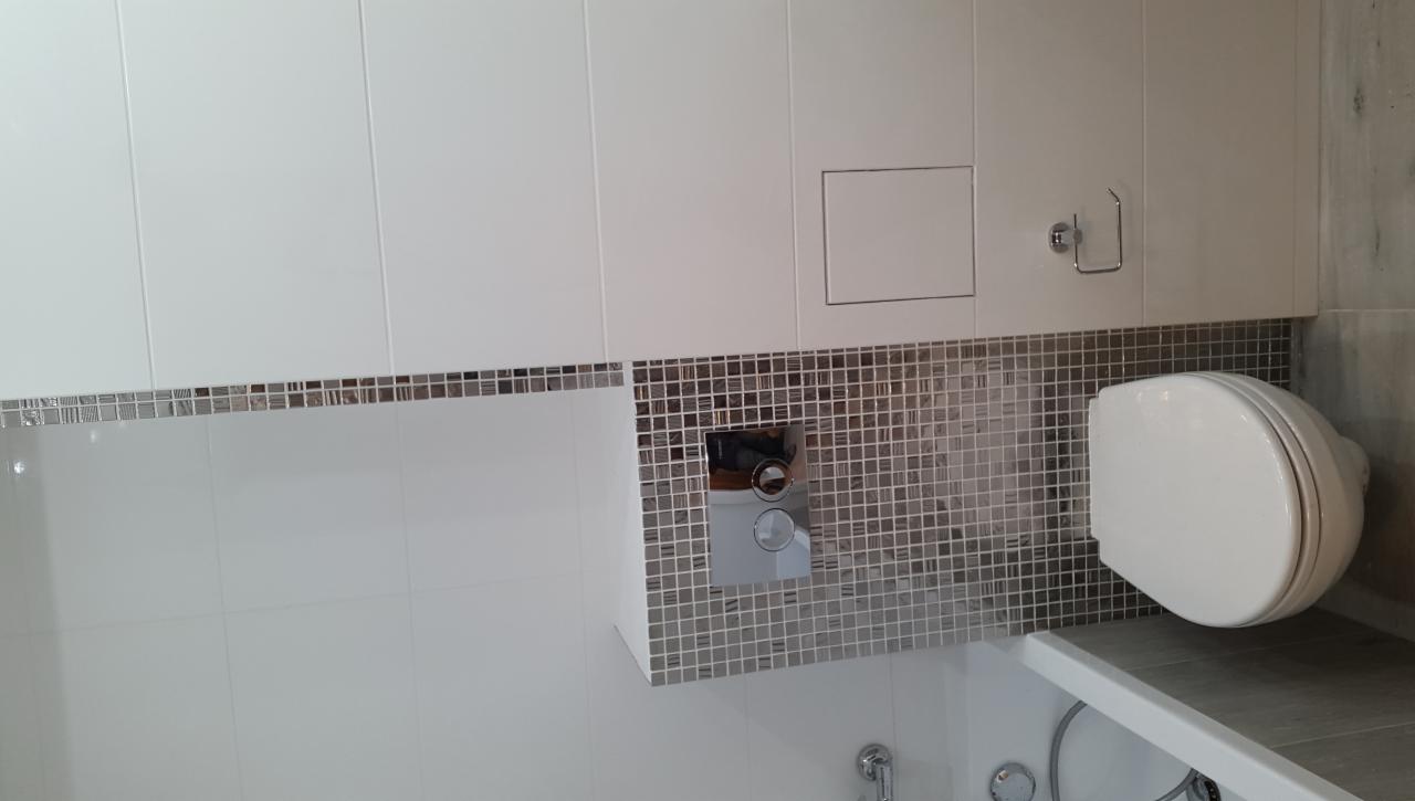 Wykończona łazienka z białą armaturą, mozaiką w odcieniach szarości i beżu, oraz ukrytym panelem dostępowym do instalacji.