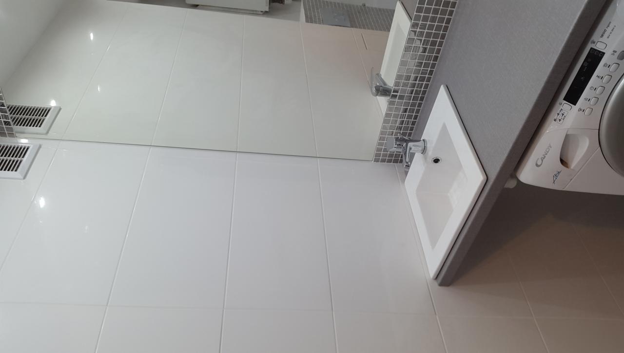 Biała łazienka z płytkami, umywalką wpuszczaną w szary blat i pralką Candy Alba, widoczne kratki wentylacyjne.