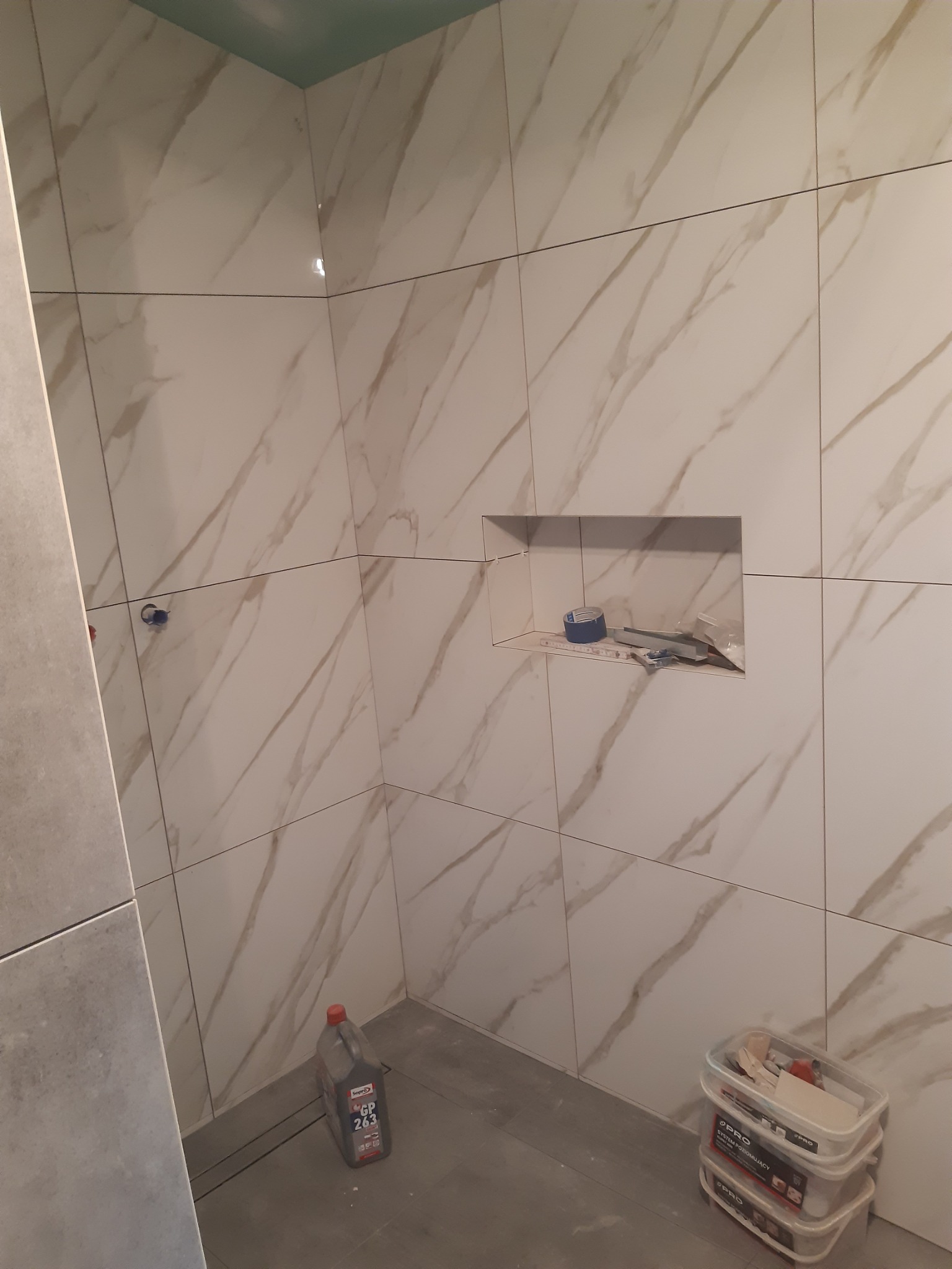Ściana w trakcie układania płytek imitujących marmur, z wnęką, w której znajdują się narzędzia, oraz pojemniki z zaprawą i płynem do czyszczenia płytek na podłodze.