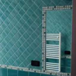 Turkusowa łazienka z ukośnie ułożonymi płytkami, dekoracyjną bordiurą i grzejnikiem drabinkowym odbijającym się w lustrze, widoczny fragment bidetu i dozownik mydła.