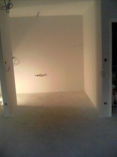 Surowe, białe wnętrze w trakcie remontu, widoczne wystające kable i puszki elektryczne w ścianie, brak podłogi, przygotowane do dalszych prac wykończeniowych.