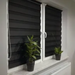 Dwa okna PCV z czarnymi roletami dzień i noc, na parapecie dwie doniczki z roślinami, widoczny biały grzejnik.