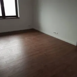 Nowo ułożona, ciemna, drewniana podłoga w pustym pokoju z białymi ścianami i oknem z ciemną ramą.