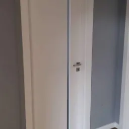 Białe drzwi wewnętrzne z ościeżnicą, widoczne na tle szarej ściany i drewnianej podłogi, z metalową klamką i zamkiem.