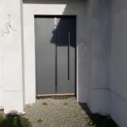 Szare drzwi wejściowe z długim, pionowym uchwytem, osadzone w surowym, białym murze z widocznymi niedoskonałościami tynku, prowadzące do wejścia wysypanego drobnym żwirem.