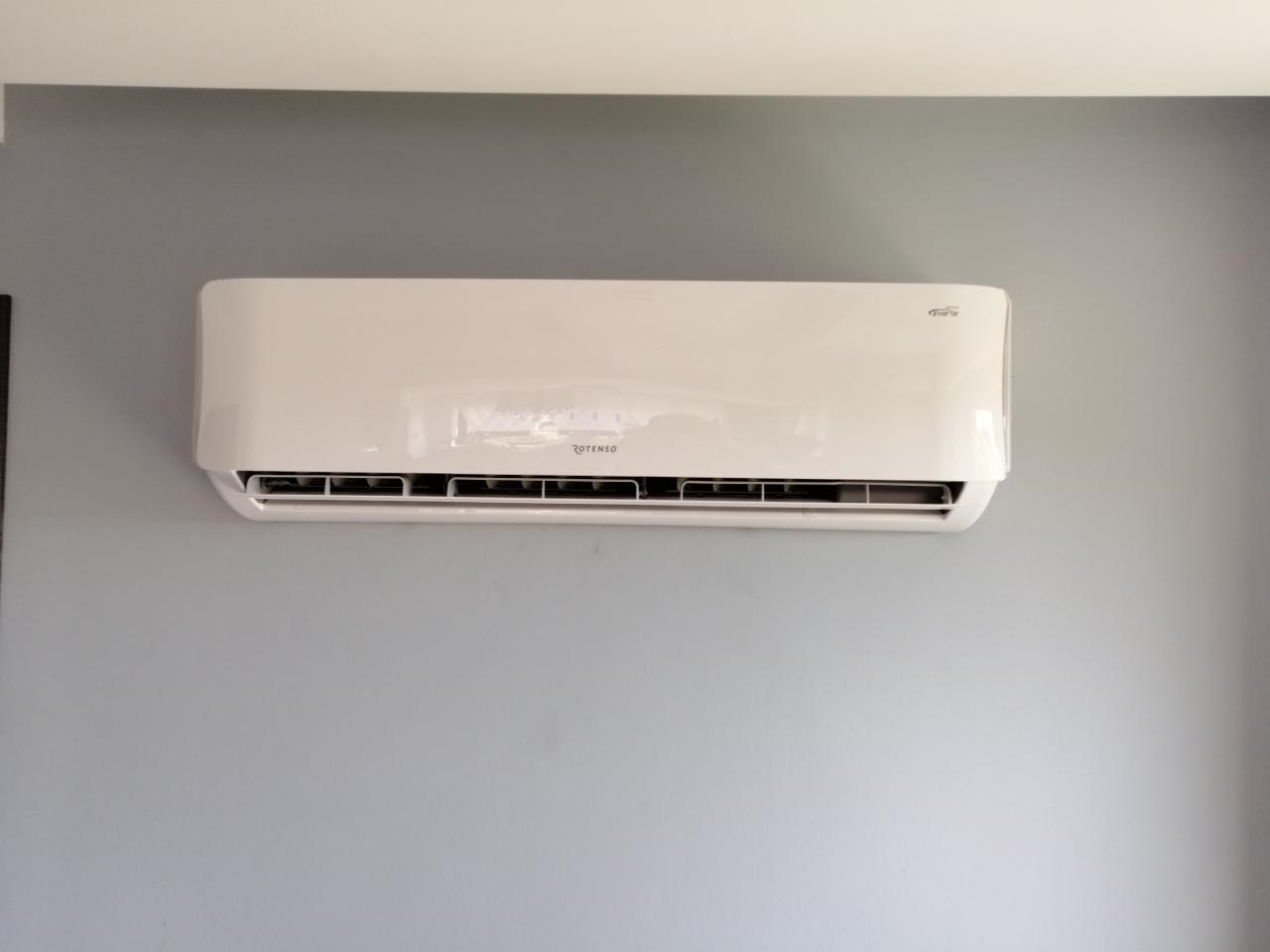 Biała jednostka klimatyzacji Rotenso Inverter zawieszona na szarej ścianie w pomieszczeniu.