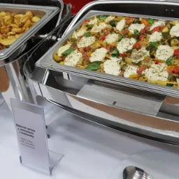 Dwa podgrzewacze bufetowe ze stali nierdzewnej zawierające dania: ziemniaki opiekane i makaron penne z warzywami i mozzarellą, oba z etykietami.