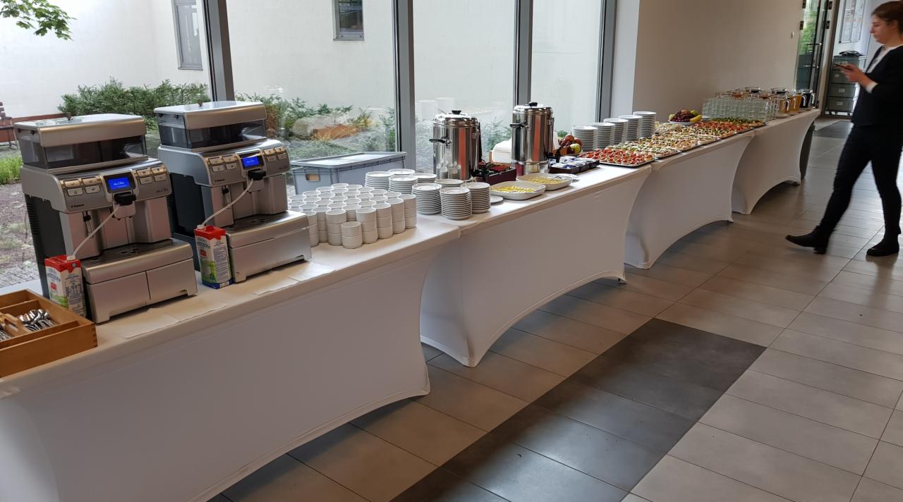 Długi stół z białym obrusem, na którym znajdują się ekspresy do kawy, stosy talerzy i filiżanek, termosy z napojami oraz przekąski; w tle okno z widokiem na zieleń i osoba w czarnym stroju.