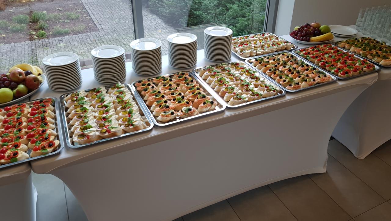 Stół pokryty białym obrusem, na którym znajdują się srebrne tace z różnorodnymi kanapeczkami, talerze w stosach i miska z owocami, ustawione przy oknie.