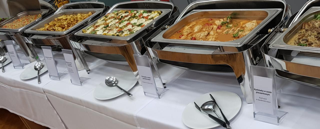 Bufet cateringowy z różnorodnymi daniami w bemarach, w tym kurczak w sosie pomidorowym i karkówka w sosie pieczarkowym, z widocznymi nazwami potraw na stojakach.