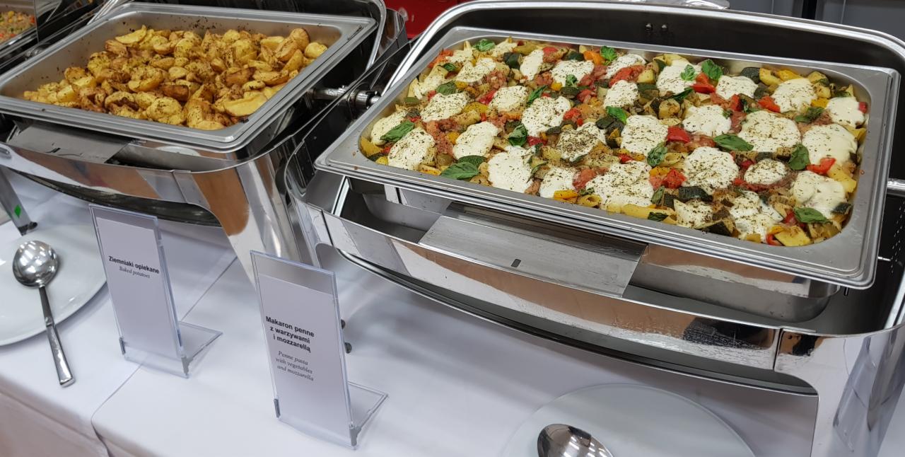 Dwa podgrzewacze bufetowe ze stali nierdzewnej zawierające dania: ziemniaki opiekane i makaron penne z warzywami i mozzarellą, oba z etykietami.