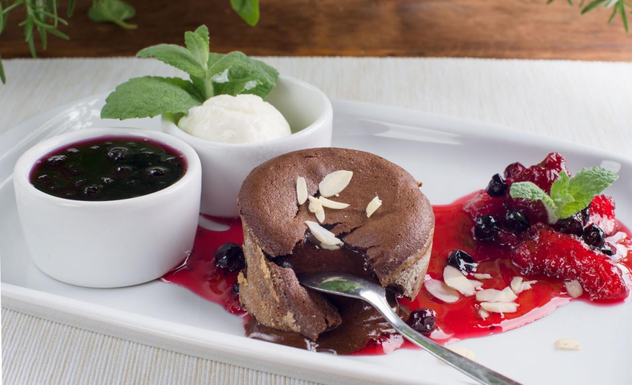 Czekoladowy fondant z płynnym środkiem, dekorowany płatkami migdałów, podany z sosem jagodowym, konfiturą z czerwonych owoców i gałką lodów na białym talerzu.