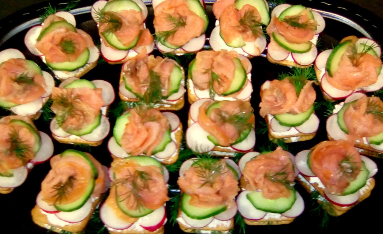 Koreczki z łososiem, ogórkiem, rzodkiewką i koperkiem na ciemnej tacy, widoczne plastry rzodkiewki z czerwoną skórką.