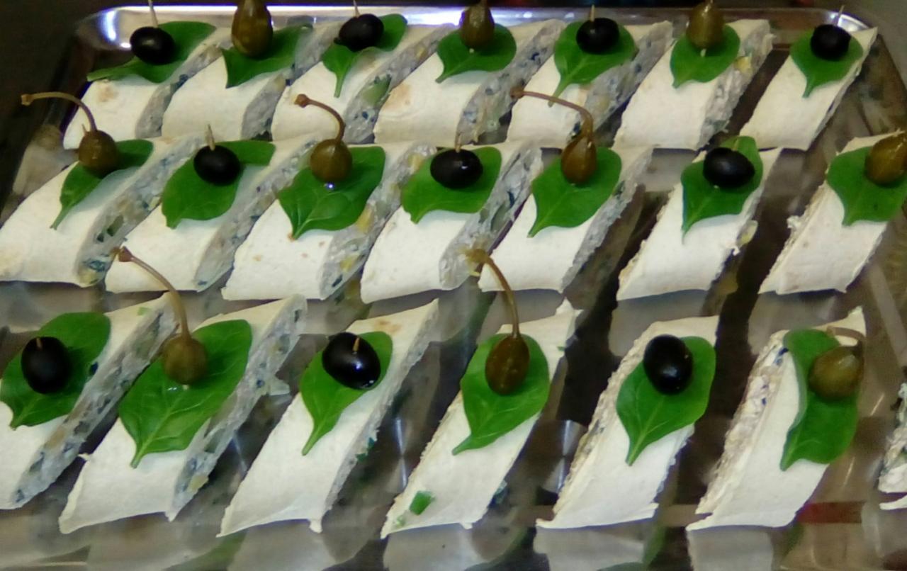 Elegancko ułożone na srebrnej tacy przekąski cateringowe we Wrocławiu: tortille z białym farszem, udekorowane zielonymi listkami bazylii, czarnymi oliwkami i kaparami.