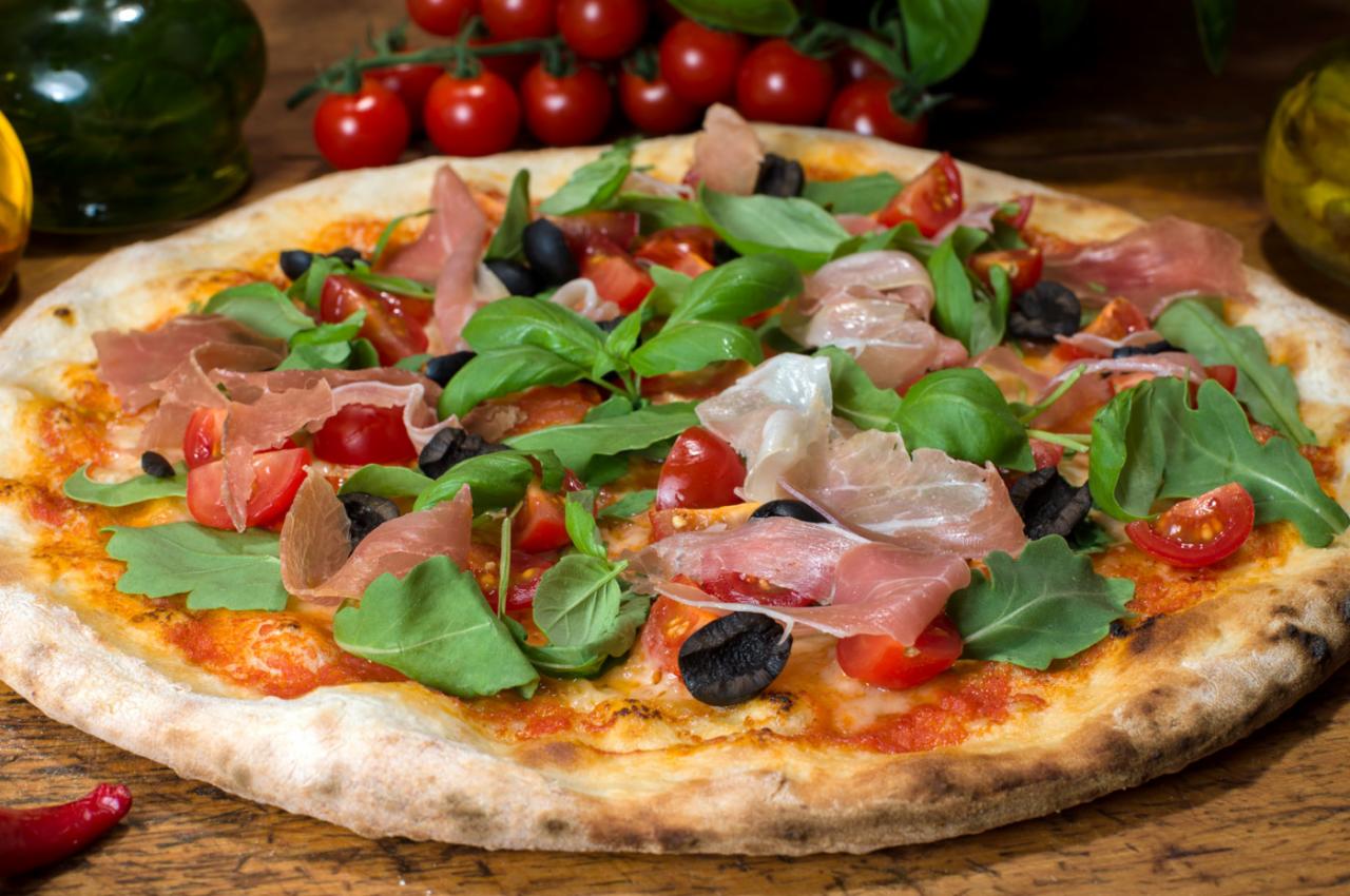 Pizza z szynką parmeńską, pomidorkami cherry, rukolą i czarnymi oliwkami na drewnianym stole.