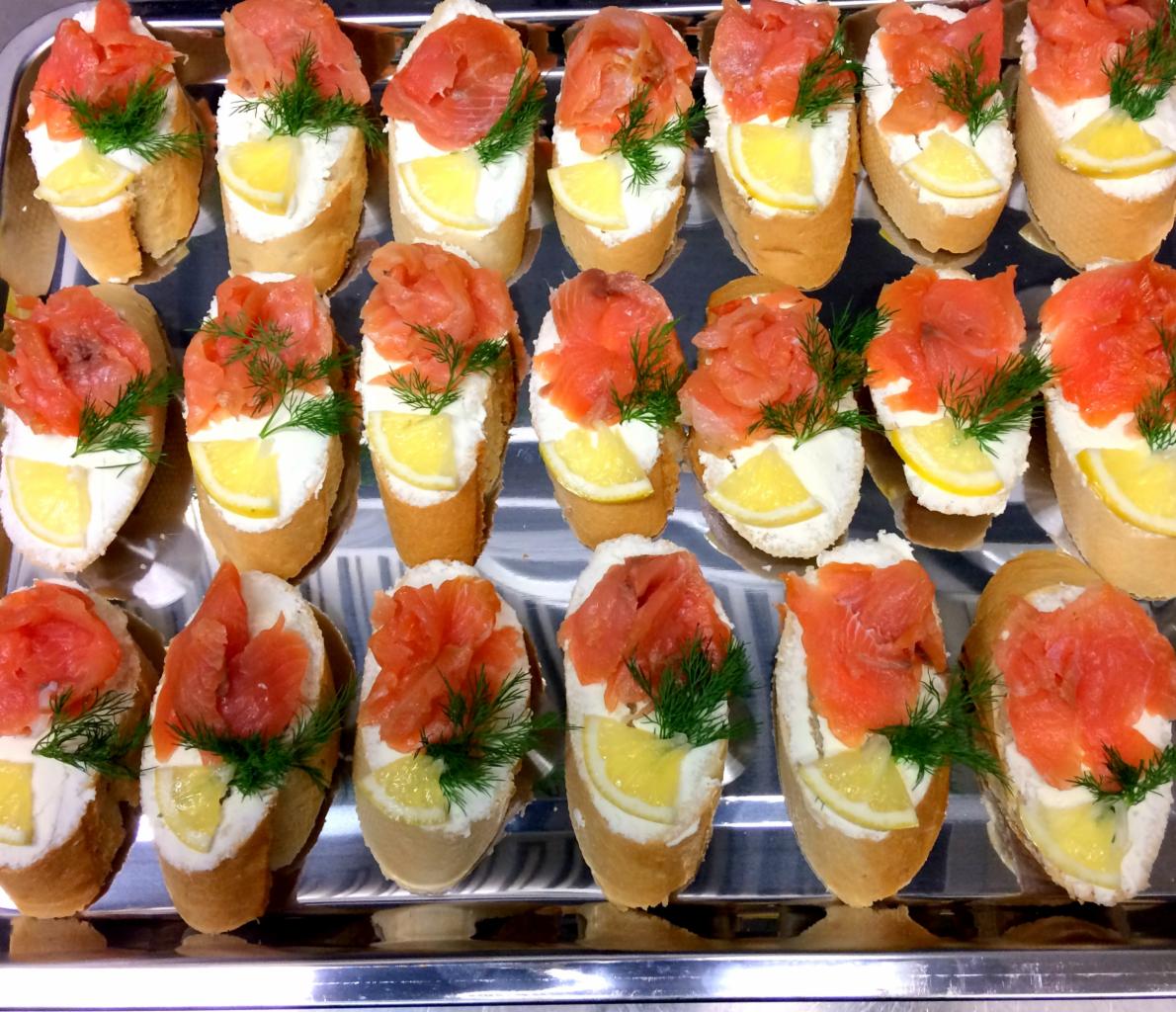 Taca z kromkami bagietki z kremowym serem, łososiem, cytryną i koperkiem, ułożonymi w równych rzędach, widoczne odbicie w metalowej powierzchni tacy.