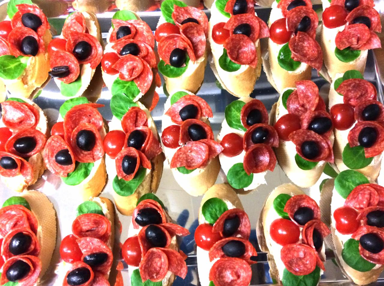 Rządki apetycznych kanapek na srebrnej tacy: bagietka z kremowym serem, listkiem szpinaku, plasterkiem salami, pomidorkiem koktajlowym i czarną oliwką.