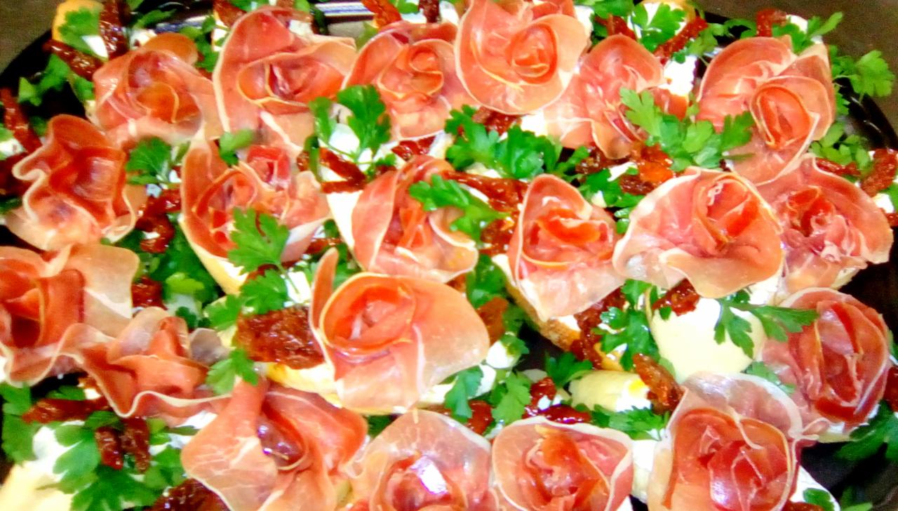 Dekoracyjne kanapeczki z szynką parmeńską ułożoną w kształt róży, suszonymi pomidorami i świeżą natką pietruszki na białym serku, podane na czarnym talerzu.