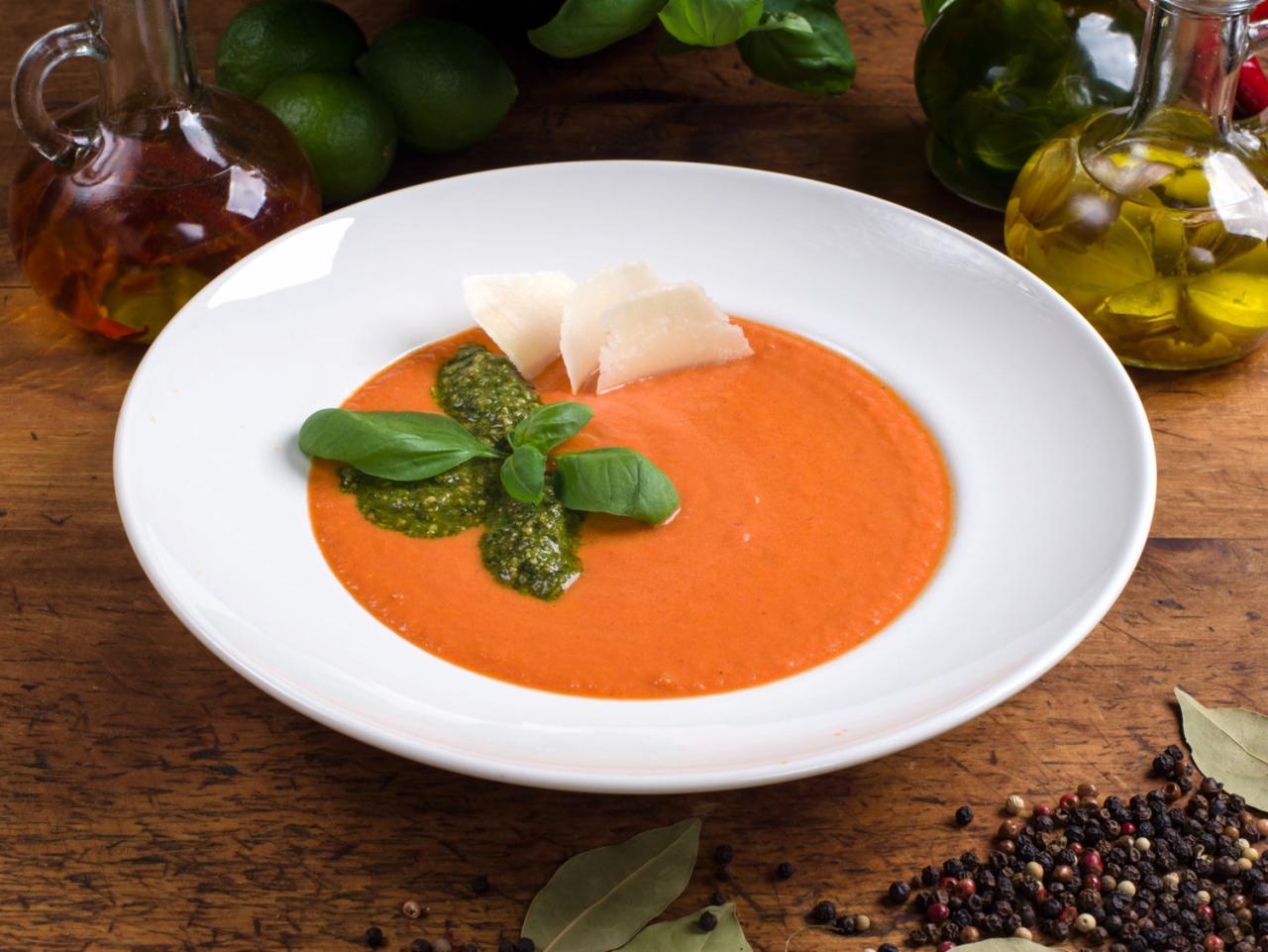 Krem z pomidorów z pesto, parmezanem i bazylią, podany na białym talerzu na drewnianym stole z przyprawami i oliwami.