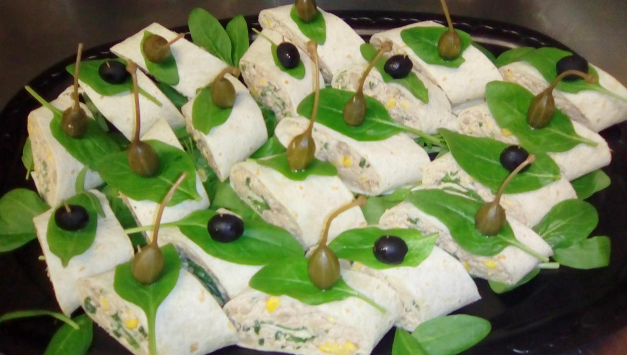 Wrapy z tortilli z farszem, dekorowane liśćmi szpinaku, kaparami i oliwkami na czarnym talerzu.