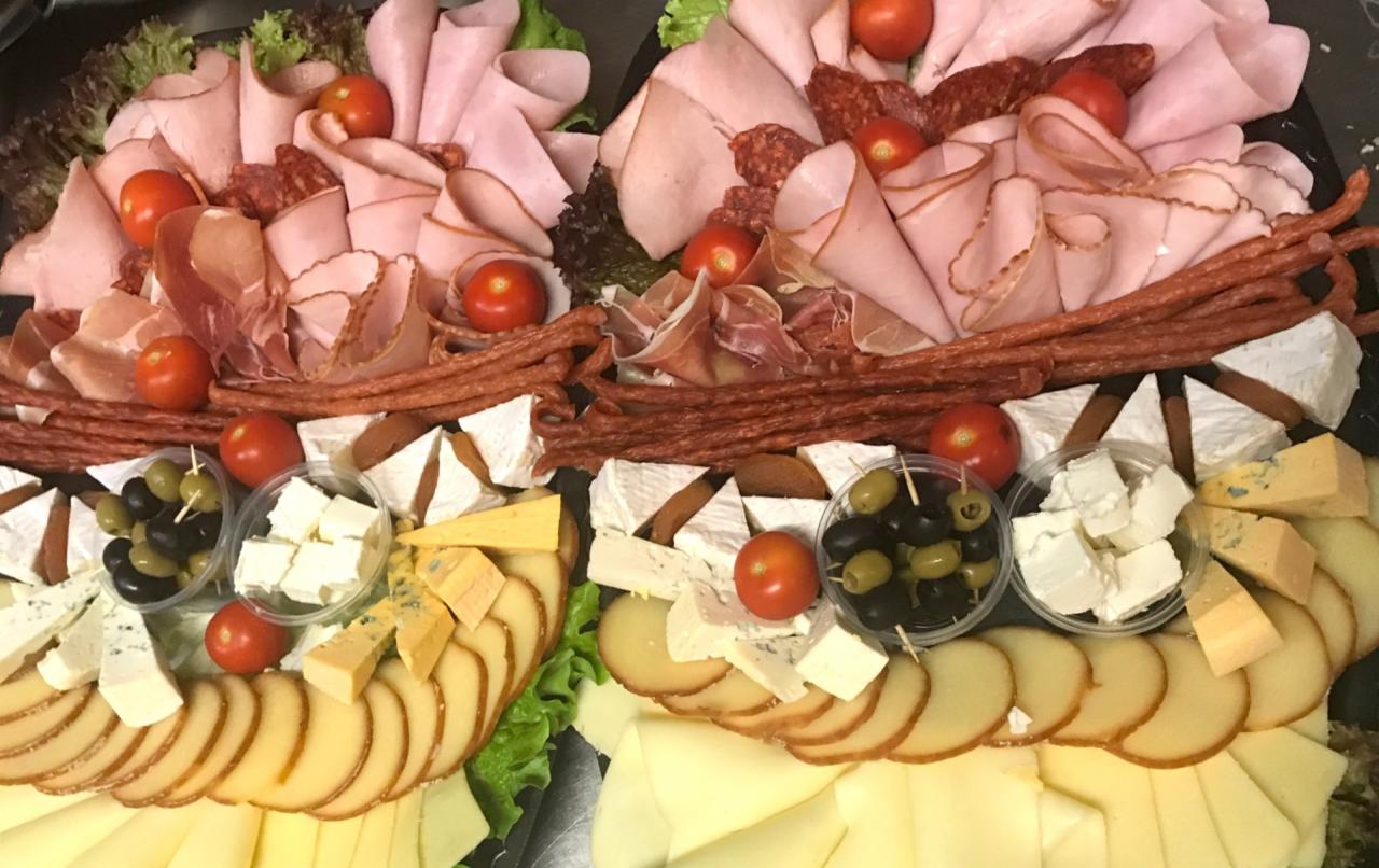 Dwie tace z selekcją wędlin, serów, oliwek i pomidorków koktajlowych, ułożonych dekoracyjnie na zielonej sałacie.