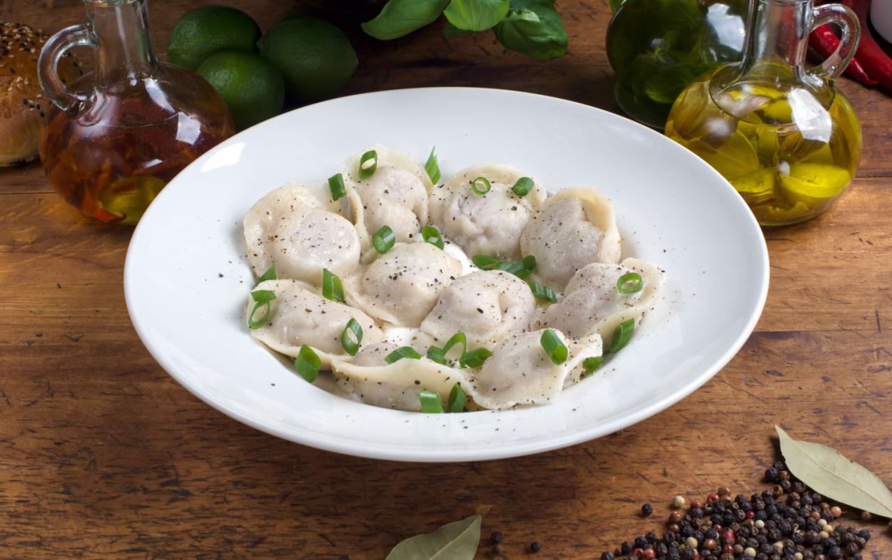 Porcja pierogów na białym talerzu, posypana pieprzem i szczypiorkiem, w tle butelki z olejem aromatyzowanym, limonki i przyprawy na drewnianym stole.