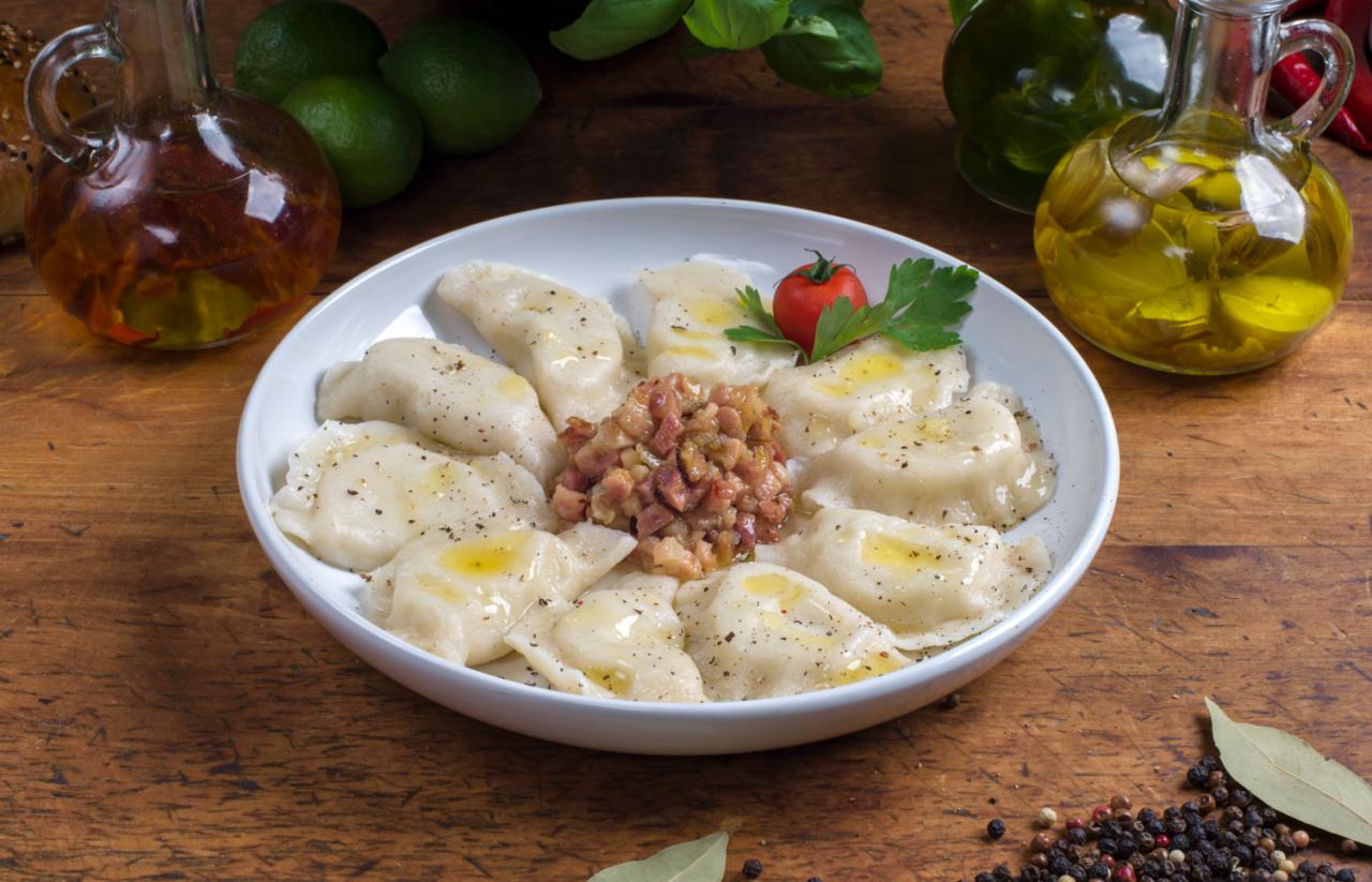 Pierogi z okrasą z boczku i olejem, podane na białym talerzu, z dodatkami w postaci świeżych ziół, limonek i przypraw na drewnianym stole.