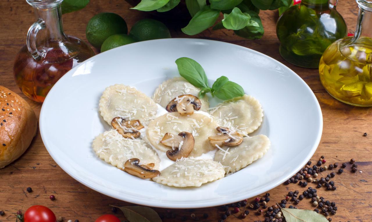 Talerz z ravioli posypanymi serem i grzybami, udekorowany bazylią, na drewnianym stole z oliwą, limonkami i pieprzem.