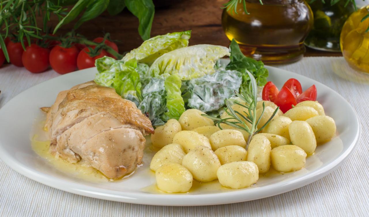 Gotowe danie na białym talerzu: pieczony filet z kurczaka, gnocchi z masłem, sałata rzymska z sosem i pomidorki koktajlowe, udekorowane gałązką rozmarynu.