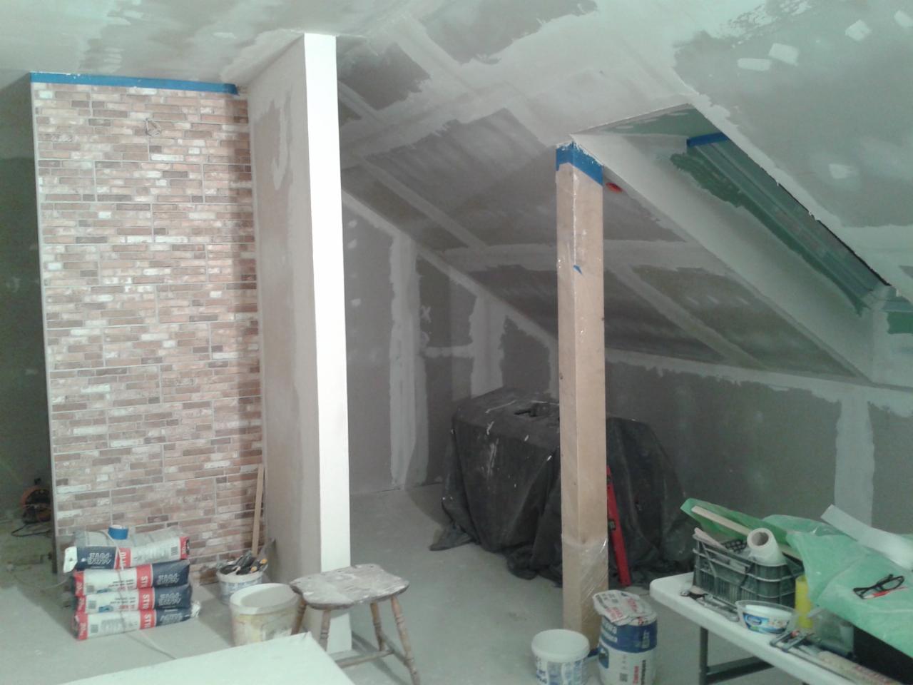 Adaptacja poddasza: ściana wykończona cegłą klinkierową, surowy stan ścian z płyt gipsowo-kartonowych, widoczne materiały budowlane i narzędzia, drewniany słupek zabezpieczony folią.