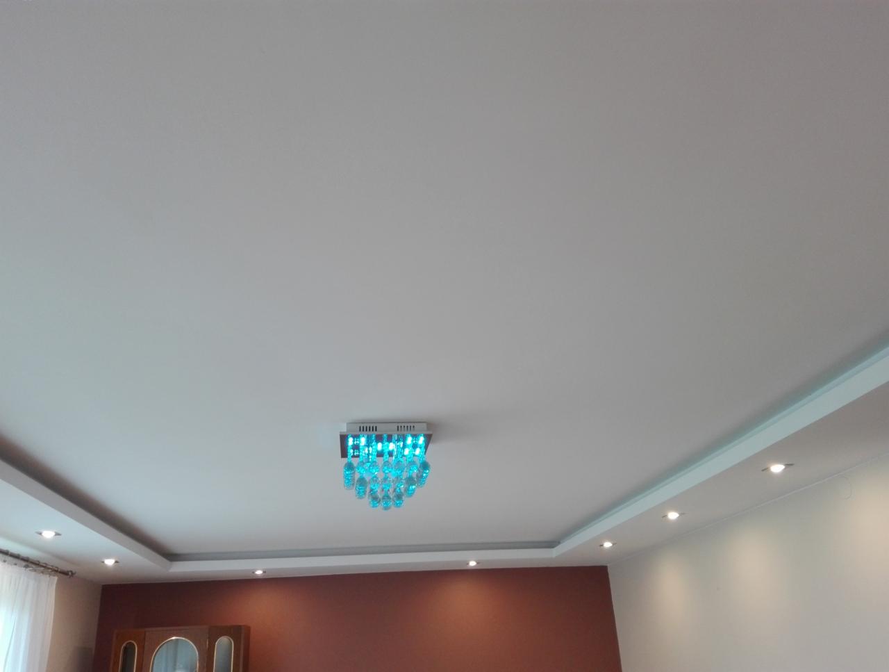 Biały sufit z dekoracyjnym oświetleniem LED w kolorze turkusowym i wpuszczanymi punktowymi lampami halogenowymi, widoczny fragment ściany pomalowanej na brązowo