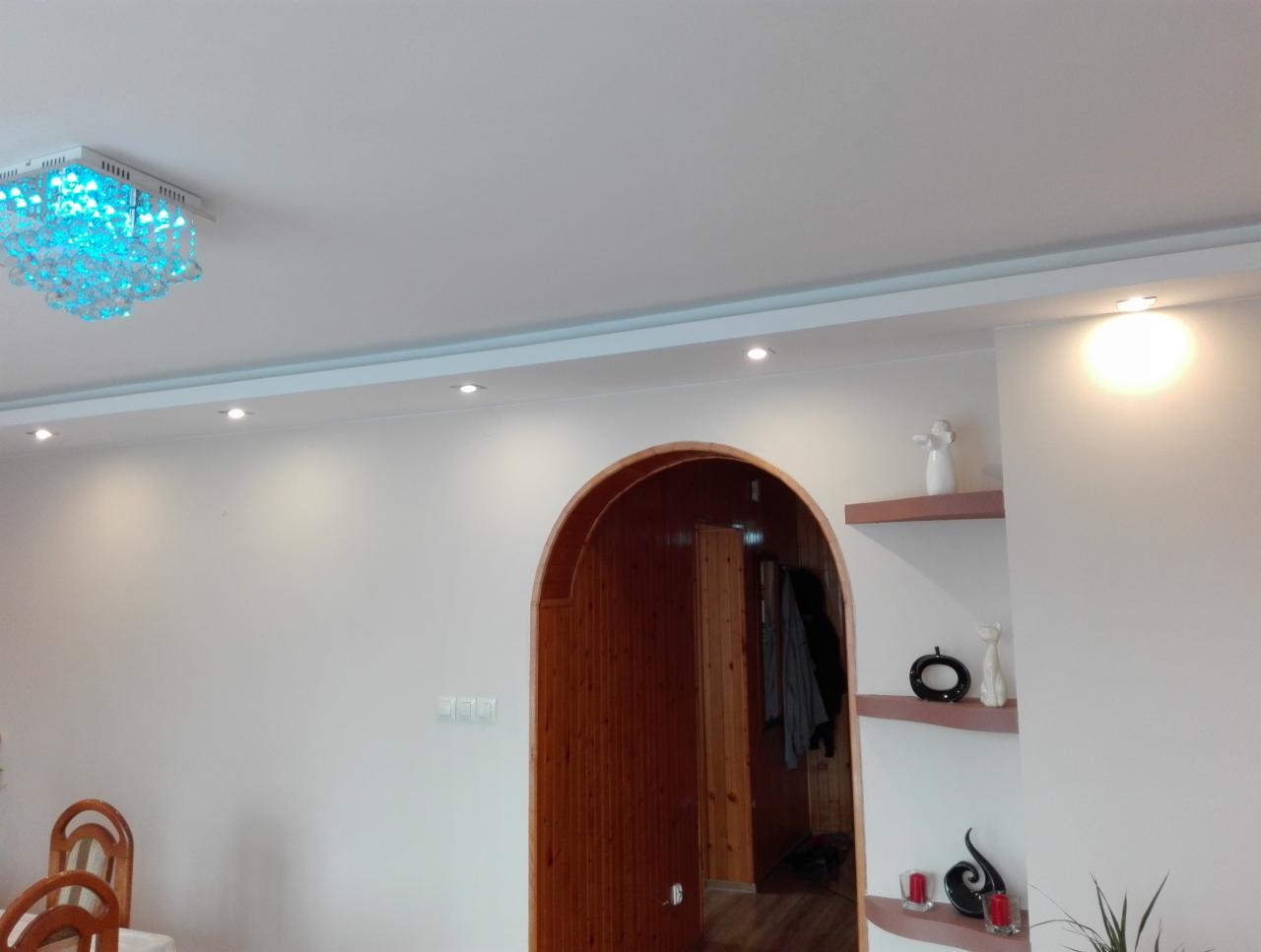 Wnętrze pokoju z łukowatym przejściem wykończonym drewnem, oświetleniem punktowym w suficie i dekoracyjnym żyrandolem z niebieskimi elementami, oraz półkami z dekoracjami.