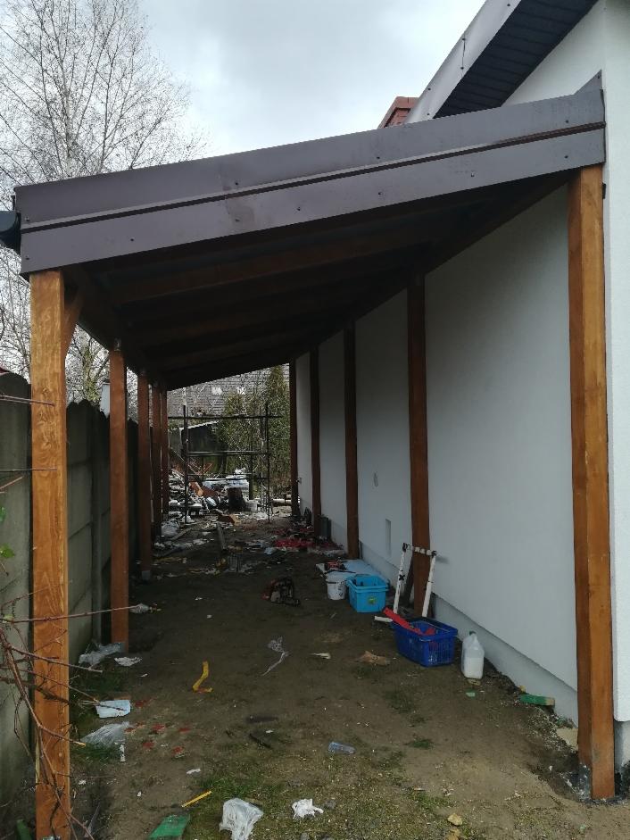 Zadaszenie tarasu z drewnianą konstrukcją i brązowym dachem, widoczne ślady prac ziemnych i materiały budowlane w otoczeniu.