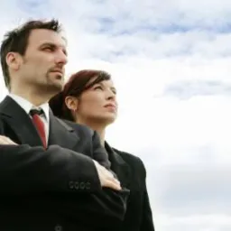 Mężczyzna w garniturze z założonymi rękami i kobieta w marynarce patrzą w górę na tle błękitnego nieba z chmurami.