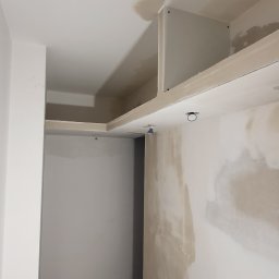 Uslugi bud - Wnętrze w trakcie prac wykończeniowych, widoczne szpachlowane ściany, zabudowa sufitowa z oświetleniem i narzędzia malarskie zabezpieczone folią.