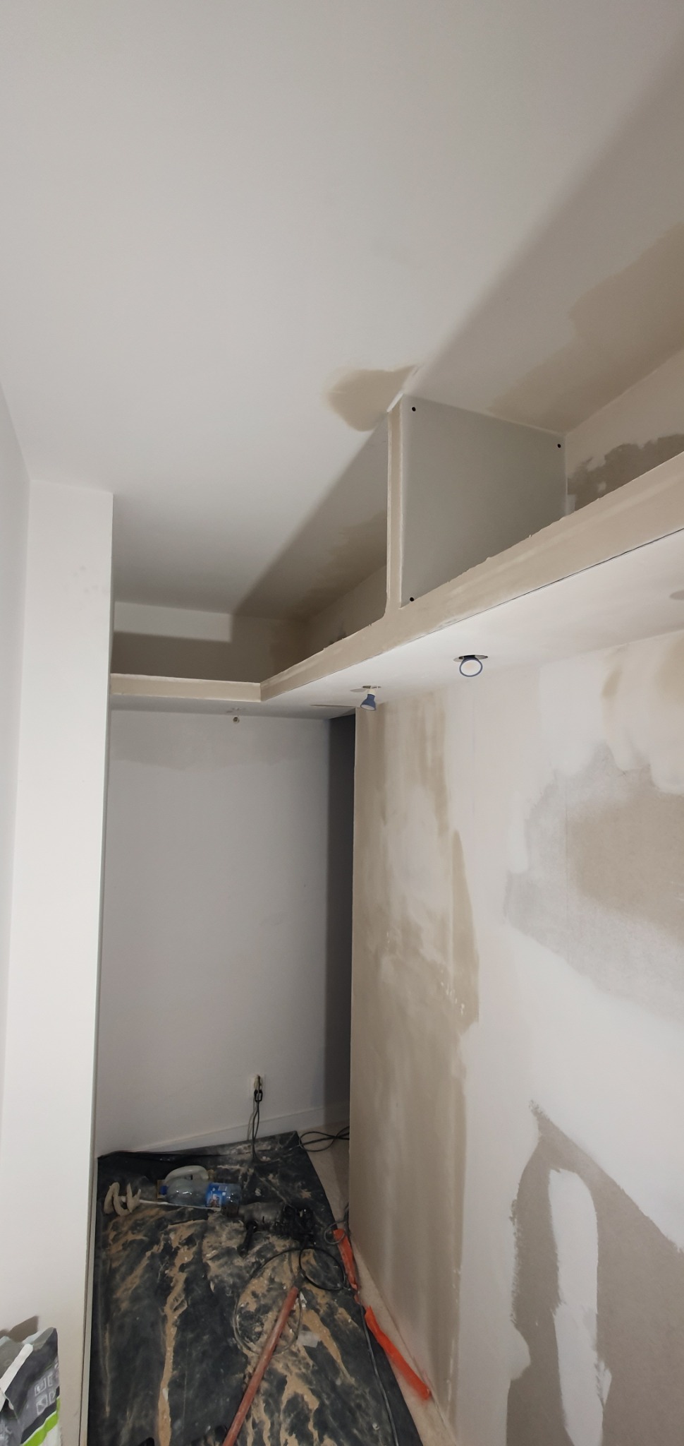 Wnętrze w trakcie prac wykończeniowych, widoczne szpachlowane ściany, zabudowa sufitowa z oświetleniem i narzędzia malarskie zabezpieczone folią.
