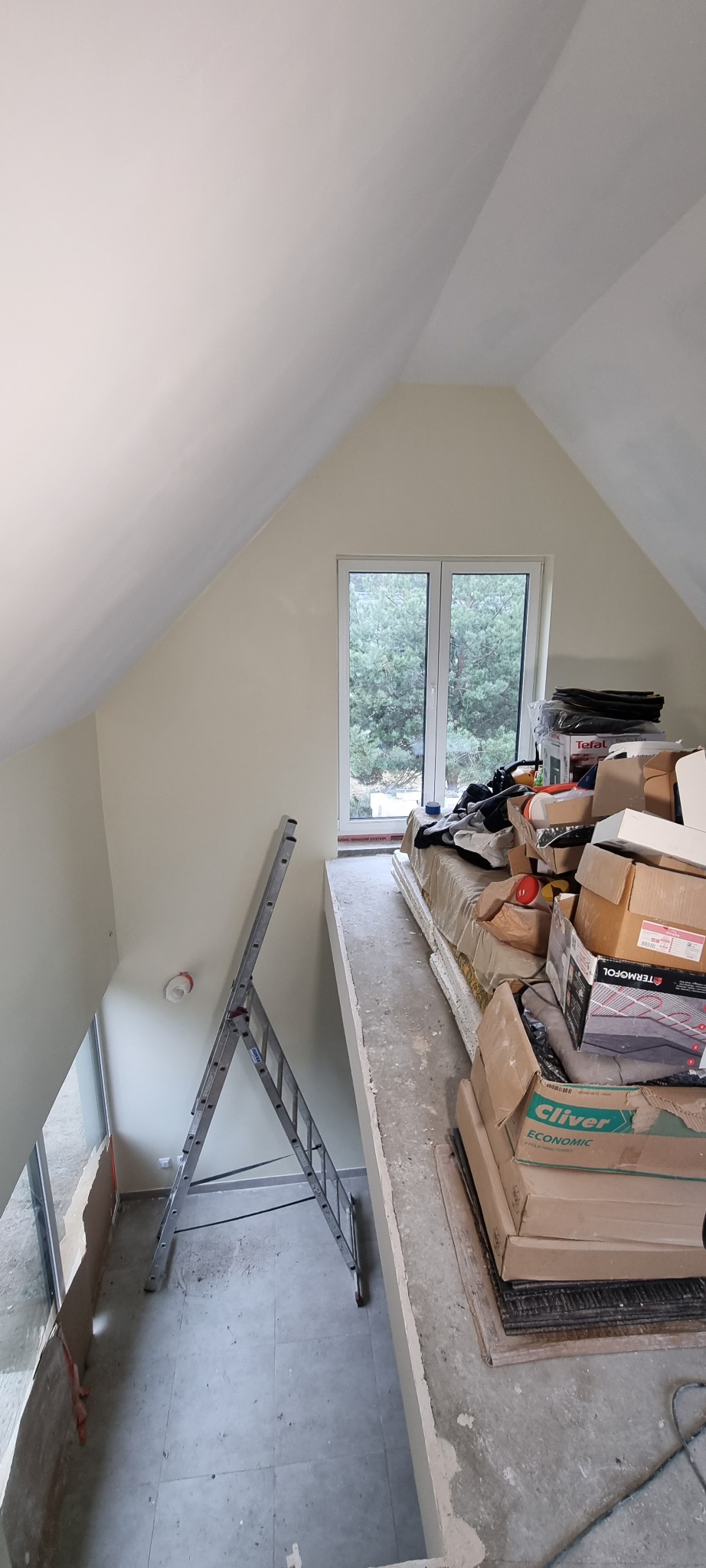 Poddasze w trakcie remontu: świeżo pomalowane ściany i sufit w jasnych odcieniach, drabina oparta o ścianę, okno z widokiem na zieleń, a na podwyższeniu sterta kartonów i materiałów budowlanych.