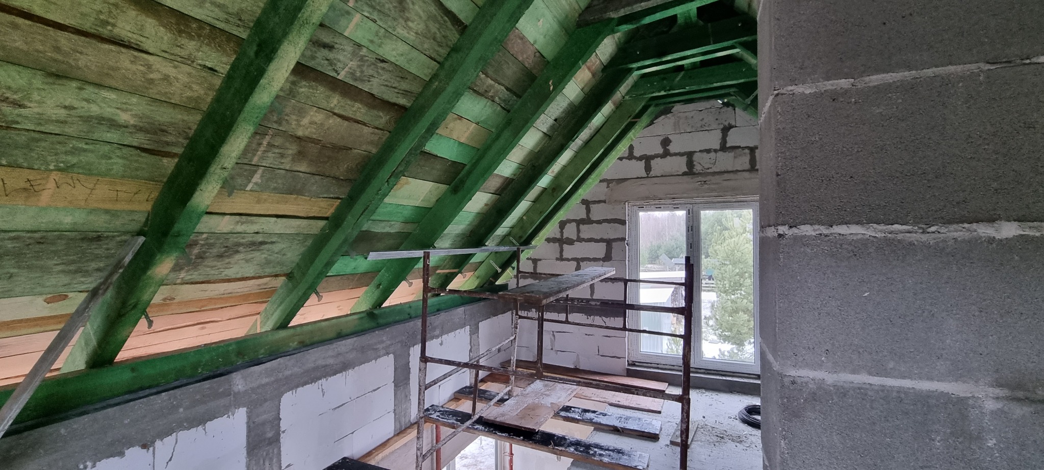 Surowe wnętrze poddasza z widoczną konstrukcją dachu pomalowaną na zielono, ścianami z bloczków betonowych, oknem i metalowym rusztowaniem.