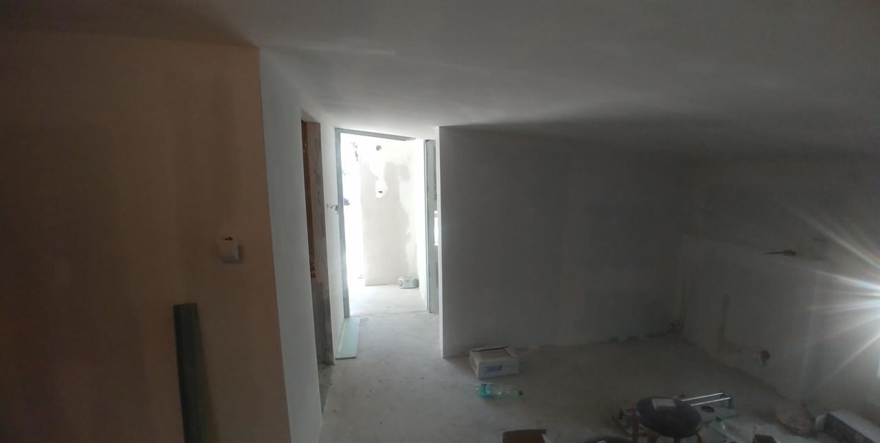 Surowe wnętrze mieszkania w trakcie prac wykończeniowych, widoczne ślady szpachlowania na ścianach i sufitach, przygotowane do dalszych etapów, z otwartym przejściem do kolejnego pomieszczenia